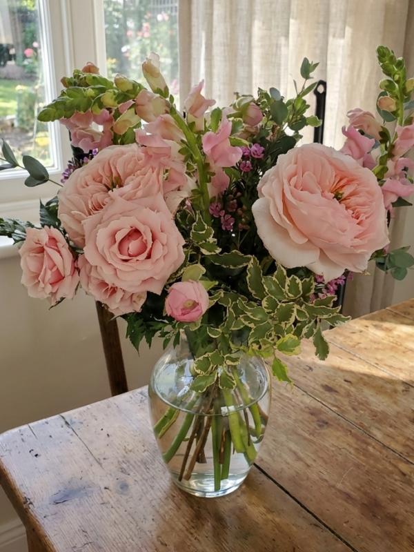 A glass vase with pink roses and assorted flowers, part of our Valentine's Day collection, sits on a wooden table near a sunlit window.