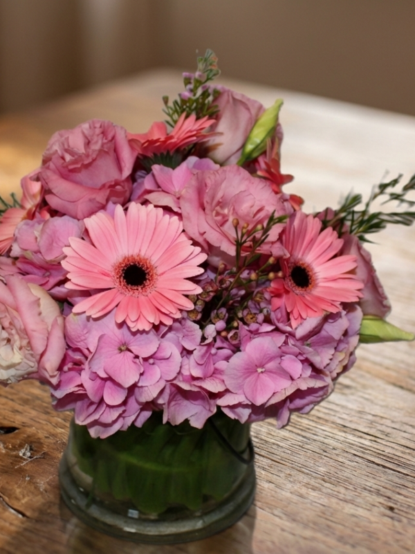 A glass vase filled with a bouquet of pink flowers sits on a wooden table.