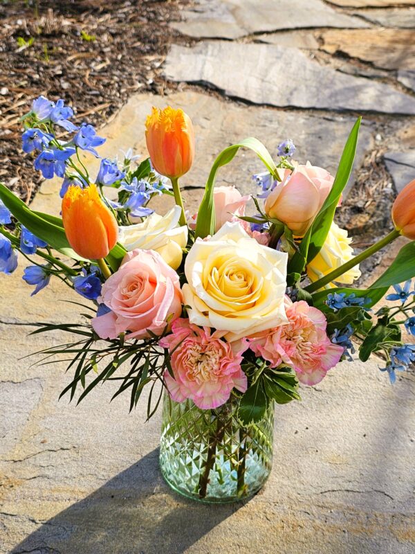 The Mother's Day Bouquet, featuring orange tulips, yellow and pink roses, pink carnations, and blue accents in a glass vase, is displayed on a sunlit stone pathway outdoors.